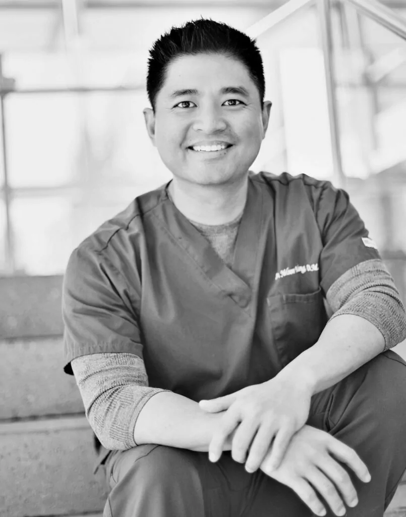 Headshot of Dr. Yang sits on a staircase wearing scrubs, often associated with medical professionals. Their arms rest casually on their knees, while the background suggests an indoor setting, possibly a clinic or hospital.