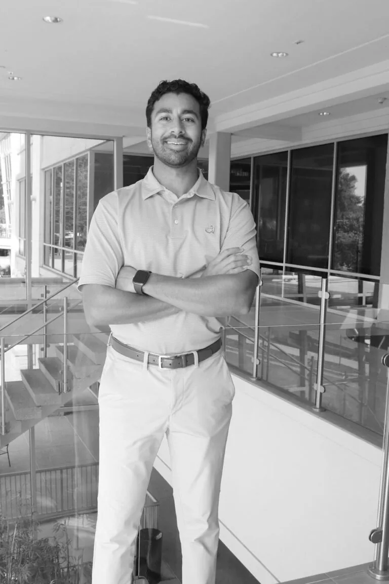 A person standing indoors, arms crossed, dressed in a light-colored polo shirt, light-colored pants, and wearing a wristwatch. The background includes glass railings, windows, and a staircase in a modern interior.