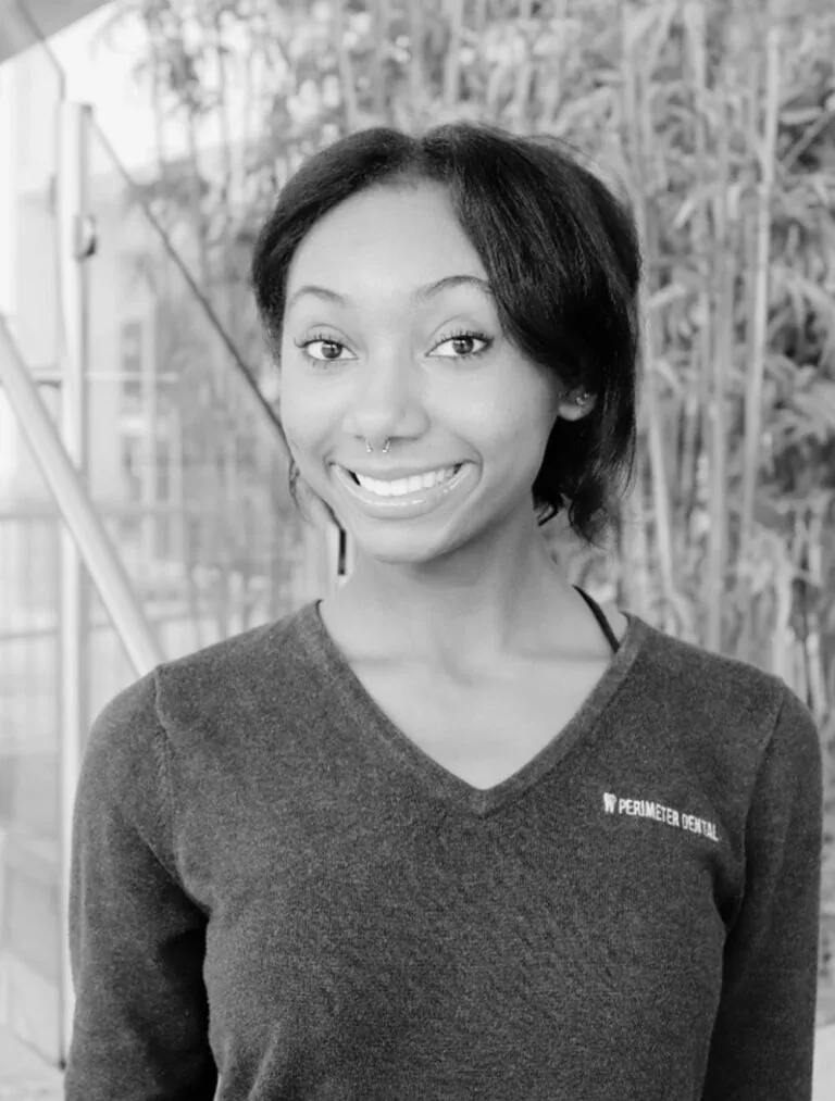 Jazlyn a dental assistant wearing a dark V-neck sweater with 'PERIMETER DENTAL' text on the left chest, standing in front of bamboo plants and a glass structure with stairs.