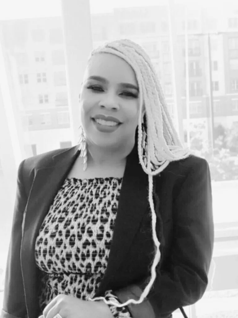 The image shows Kirah an administrator, dressed in a patterned dress paired with a dark blazer. Their long, braided hair is light in color. The background reveals an urban scene with visible buildings and windows.