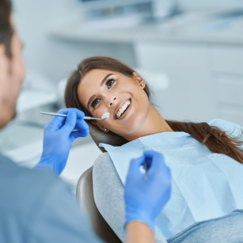 A dentist in blue gloves performing a dental check-up on a patient in a reclining chair, with a blue dental bib and precision instruments in hand.